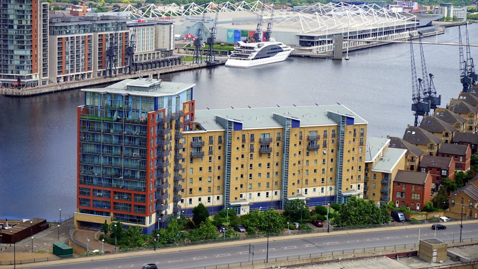 A cluster of modern high-rise apartment buildings with large glass balconies and colorful facades overlooking a river in South Quay Docklands. In the background, there is a marina with white yachts do