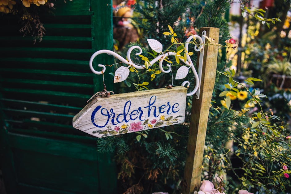 A rustic wooden sign with the words 'Order here' painted in blue cursive lettering, hanging from a decorative white iron bracket with scrollwork at the top. The sign is attached to a wooden post amids