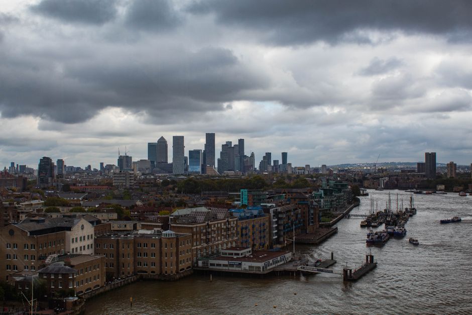 A wide view of Canary Wharf skyline under dark, cloudy skies, featuring tall glass skyscrapers and modern office buildings in the distance, with a river in the foreground where boats are moored along