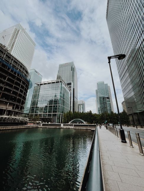 This image depicts a modern cityscape in Canary Wharf with tall glass skyscrapers reflecting the sky and clouds. In the foreground, there is a waterway with calm, greenish water, bordered by a paved w
