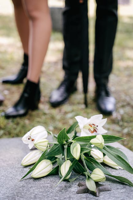 A delicate floral arrangement of white lilies and closed lily buds with green leaves, arranged as a floral gift or tribute, resting on a stone surface outdoors. The lilies are fresh, with smooth white